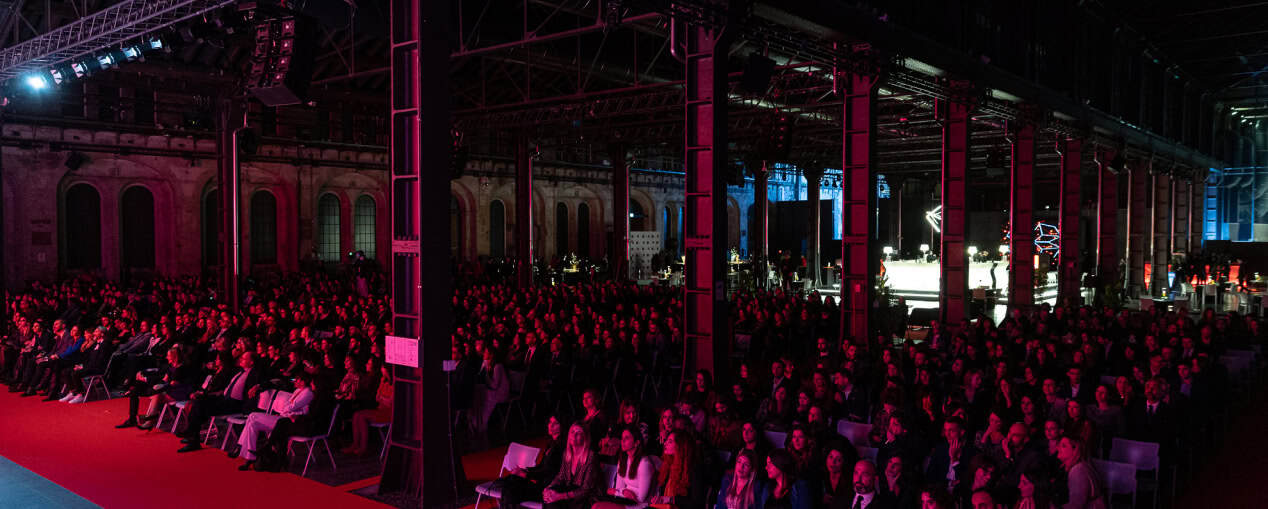 Pubblico numeroso seduto in un evento indoor con illuminazione rossa e strutture metalliche.